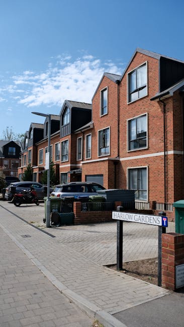 Image shows a multi-storey residential building with a modern design, constructed with red brickwork and featuring large rectangular windows, some with black window frames. The building is situated along a paved street with parked cars, including a black SUV and a silver vehicle, visible in front of the property. Near the sidewalk, a small black metal fence surrounds a garden area with a green rubbish bin beside it. A street sign reading 'Harlow Gardens' is mounted on a black post at the corner of the pavement, indicating the location. The sky above is bright and partly cloudy, casting natural daylight onto the scene. This urban residential setting, typical of a housing development, is suitable for house removal and relocation services, with clear access for moving trucks and equipment.