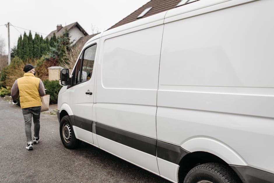 A man wearing a black cap, face mask, beige vest over a grey long-sleeve shirt, and dark grey trousers is standing on a paved driveway in front of a white commercial van. He is holding the van’s sid