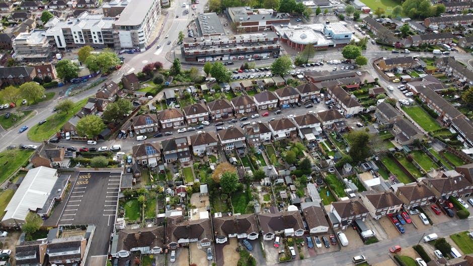 An aerial view of a residential neighbourhood showing multiple terraced houses with small gardens, some featuring patio furniture and decorative plants. Driveways and parked cars line the street, while a larger commercial or apartment complex is visible in the background. In the foreground, a parking lot adjacent to a commercial building is seen, with visible markings and parked vehicles. The area is surrounded by trees and green spaces, with a mix of urban and suburban elements. The lighting appears to be natural daylight. This setting illustrates the type of environment where house removals and furniture transport services provided by Man with Van Harlington might take place, highlighting typical packing, loading, and logistics processes involved in local home relocation.