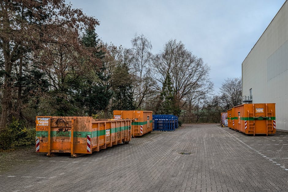 A paved outdoor area adjacent to a large white industrial building, featuring several orange metal skips with green and white reflective stripes lined up along the edge of the paved surface. The skips are for waste disposal and are marked with signage indicating disposal company details, with some showing signs of previous use and dirt marks. Behind the skips, there are leafless trees and some evergreen trees in the background, suggesting a late autumn or winter setting. The outside space is illuminated by natural daylight under a cloudy sky, and the area appears to be part of a home relocation or moving logistics process, where waste materials and packing debris are collected before or after furniture transport. The scene is captured in a manner that emphasizes the organization of waste management during a move, fitting within the context of professional removals by Man with Van Harlington.