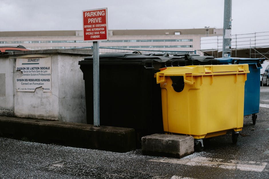 A paved outdoor area adjacent to a large white industrial building, featuring several orange metal skips with green and white reflective stripes lined up along the edge of the paved surface. The skips are for waste disposal and are marked with signage indicating disposal company details, with some showing signs of previous use and dirt marks. Behind the skips, there are leafless trees and some evergreen trees in the background, suggesting a late autumn or winter setting. The outside space is illuminated by natural daylight under a cloudy sky, and the area appears to be part of a home relocation or moving logistics process, where waste materials and packing debris are collected before or after furniture transport. The scene is captured in a manner that emphasizes the organization of waste management during a move, fitting within the context of professional removals by Man with Van Harlington.