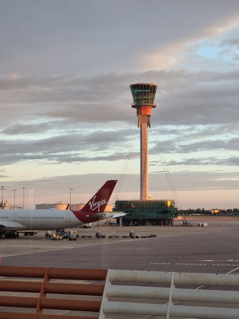 An airport tarmac scene showing a Virgin Atlantic aircraft parked near the terminal, with its distinctive red tail fin and logo visible. In the background, a control tower with a cylindrical shape and antennas at the top stands against a partly cloudy sky at sunset, casting a warm glow. The foreground features part of a boarding gate with metal railings and orange structural elements, as well as ground support equipment like baggage carts. The overall scene captures the busy environment of an international airport, illustrating the logistics involved in home relocation and furniture transport services, supported by Man with Van Harlington’s removals team, near Heathrow.