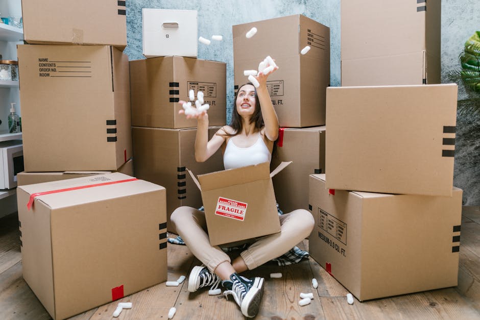 A young woman sitting on a wooden floor surrounded by numerous brown cardboard moving boxes of various sizes, some sealed with red tape, and others open with packing foam peanuts spilling out. She is inside a room with a textured light blue wall and a part of a potted plant visible on the right. The woman is casually dressed in a white sleeveless top, beige pants, and black sneakers, smiling as she throws packing foam peanuts into the air. Several cardboard boxes are stacked behind her, with some placed directly on the floor and others arranged in a bin-like manner. There is a white microwave on a countertop in the background, indicating a domestic setting prepared for a home relocation or moving process. Man with Van Harlington specializes in professional removals, including packing, loading, and furniture transport, as reflected in the scene illustrating the packing and moving stages.