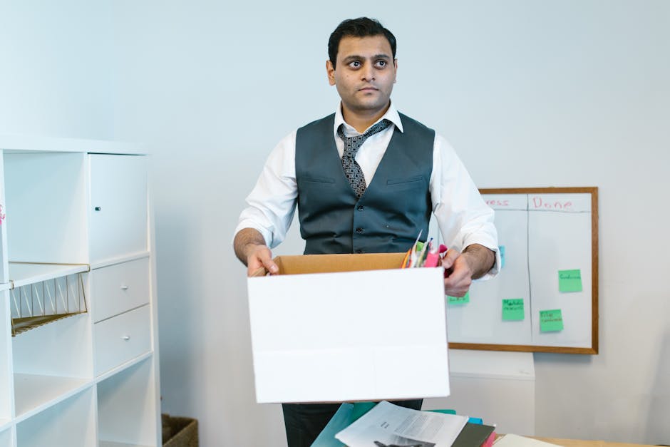 A man with Van Harlington, dressed in a white shirt, dark vest, and patterned tie, is inside an office space holding an open cardboard box filled with various office supplies, including pens and paper