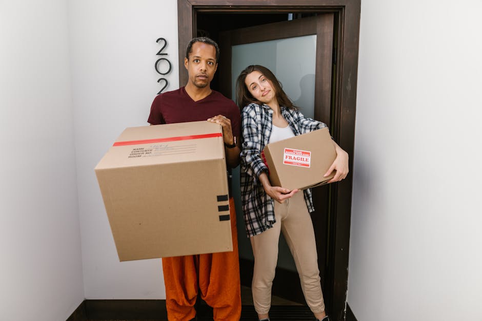 A man and a woman stand inside a building's doorway, each holding a cardboard box during a home relocation process. The man, dressed in a maroon shirt and orange pants, holds a large box with red and black tape, while the woman, wearing a plaid shirt and beige pants, holds a smaller, labeled fragile box. They are positioned directly in front of a dark brown doorframe with a frosted glass panel, and there are visible house number numbers on the wall beside them. The environment is well-lit, with a plain light-colored wall on one side and a darker floor beneath their feet. Behind them, inside the interior, there are no visible furnishings or additional items, suggesting they are in the process of loading or unloading furniture and boxes during a house move. The image reflects the logistics and careful handling involved in packing and moving, aligning with services offered by Man with Van Harlington, known for same-day removals and last-minute booking solutions.