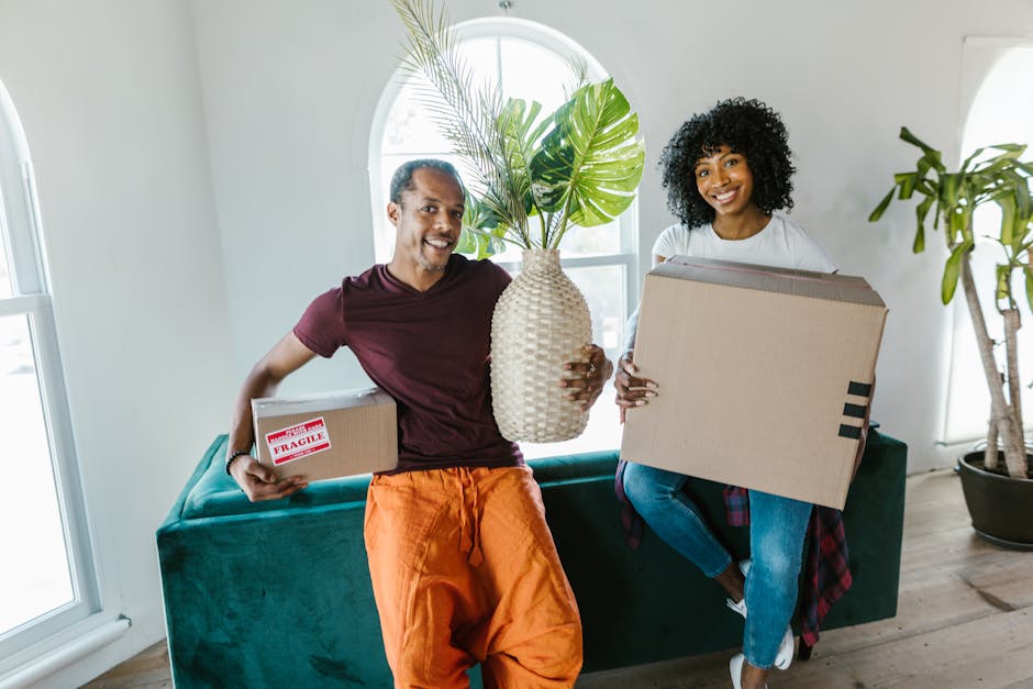 A man and a woman stand inside a building's doorway, each holding a cardboard box during a home relocation process. The man, dressed in a maroon shirt and orange pants, holds a large box with red and black tape, while the woman, wearing a plaid shirt and beige pants, holds a smaller, labeled fragile box. They are positioned directly in front of a dark brown doorframe with a frosted glass panel, and there are visible house number numbers on the wall beside them. The environment is well-lit, with a plain light-colored wall on one side and a darker floor beneath their feet. Behind them, inside the interior, there are no visible furnishings or additional items, suggesting they are in the process of loading or unloading furniture and boxes during a house move. The image reflects the logistics and careful handling involved in packing and moving, aligning with services offered by Man with Van Harlington, known for same-day removals and last-minute booking solutions.