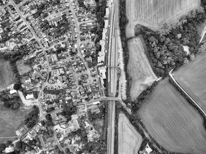 An aerial black and white photograph shows a densely populated residential area with numerous houses, gardens, and streets on the left side, transitioning to open fields and patches of woodland on the right. A railway line runs vertically through the center, with railway tracks and a station platform visible. Adjacent to the railway, there is a car park with parked vehicles and a bridge crossing over the tracks, connecting the two sides of the neighborhood. The image captures the urban and rural boundary, illustrating the layout of homes, roads, and green spaces, which could relate to house removals or home relocation planning, as offered by Man with Van Harlington for efficient furniture transport and packing during moves.