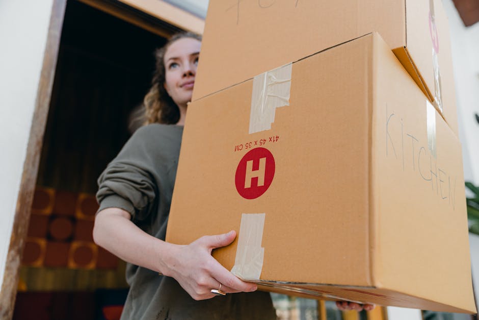 A young woman with long, wavy brown hair wearing a grey sweater is inside a home, carrying a large cardboard box marked with a red 'H' and printed dimensions, positioned near the building's doorway. She is holding the box securely with both hands, preparing for a home relocation or furniture transport process. The box is sealed with beige packing tape and has handwritten labels on it, indicating it contains household items for packing and moving. In the background, there is a partially open door revealing a storage area or room with stacked cylindrical items. The interior lighting is moderate, and the floor appears to be a neutral tone. The scene is part of a professional house removals service, such as Man with Van Harlington, which handles packing, loading, and transportation as part of the moving logistics.