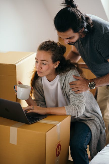 A person wearing a grey knitted sweater is seen holding a large cardboard moving box with clear packing tape sealing the top flaps, inside a home environment. The box displays a red 'H' logo and is positioned in front of a white sofa with soft cushions. Behind the person, additional packed boxes are visible on the wooden floor, some closed and taped, indicating preparation for a home relocation or furniture transport. The background includes a blurred green plant, suggesting natural indoor lighting. The scene captures the process of packing and loading items, reflecting professional house removals services by Man with Van Harlington, as part of a quick and secure moving operation for students or residents. The setting emphasizes the careful handling of household items during packing, highlighting the logistics involved in home removals and move-outs.